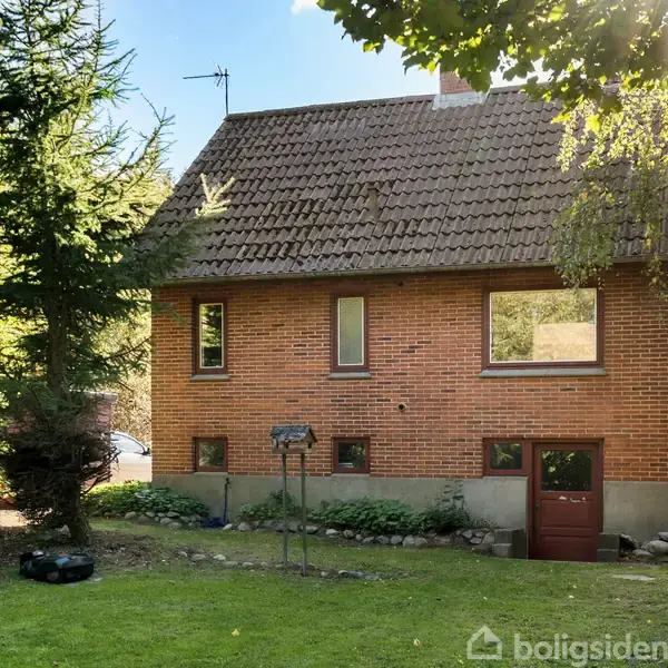 Et rødt murstenshus står med et skråt tag og flere vinduer, omgivet af en grøn have med græs og træer. Sollys skinner gennem løvet.