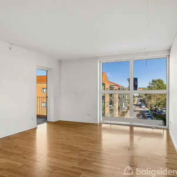 Lejlighedskompleks med altaner vender ud mod en grøn gård og parkeringsplads. Omkringliggende bygninger ses under en klar blå himmel.
