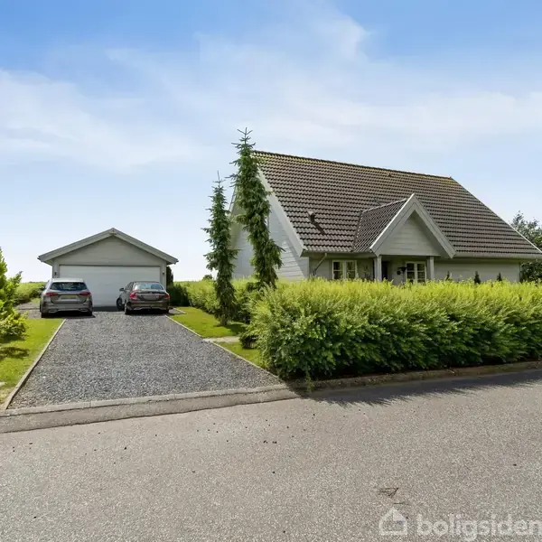 En hvid villa med gråt tag står i en grøn have ved siden af en indkørsel med to biler. En separat garage ses i baggrunden under en klar blå himmel.
