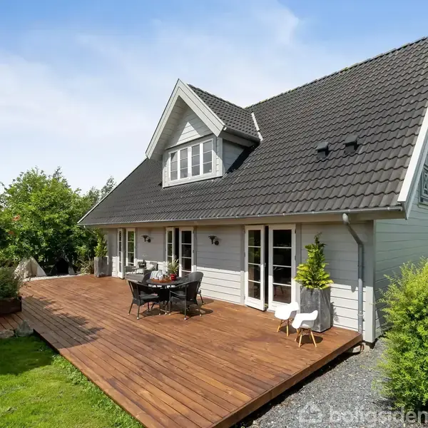 Et hus med mørkt tag og lyse vægge har en træterrasse med havemøbler foran, omgivet af en have med græs og buske under en klar himmel.