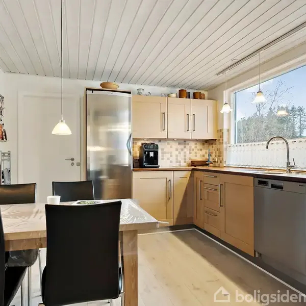 Et køkken med træskabe og metalhåndtag, hvor en kaffemaskine står på bordet. Ved siden af er et stort vindue, og et spisebord med fire sort-stole er i forgrunden.