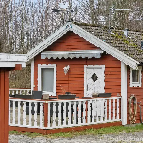 Rødt træhus med hvidmalet terrasse og dør, beliggende i en skovklædt område. Et skilt med tallet "2" ses på et mindre skur.