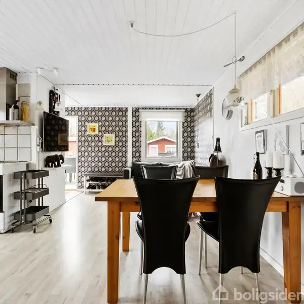 Spisebord med sorte stole står i et lyst køkken-alrum med hvide vægge og træloft. Et mønstret tapet pryder væggen, og et vindue afslører en rød bygning udenfor.