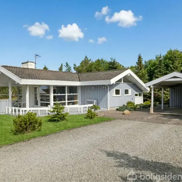 Et gråt sommerhus med hvidt tag står på en grusplads omgivet af grønne træer og buske under en blå himmel med få skyer.