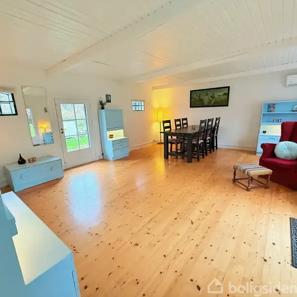 Stue med træbord og seks stole, rødt lænestol og fodskammel. Blå skabe og en hylde langs væggen. Lyse trægulve, hvidt loft og vægge, dør til haven.