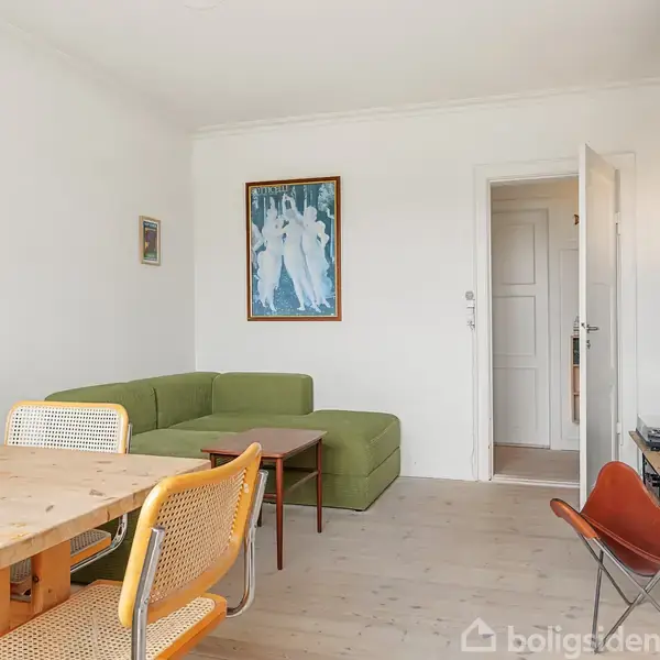 Grønt hjørnesofa sæt står i en lys stue med hvide vægge. Langt træbord omgivet af stole, maleri på væg og åben dør til et andet rum. Hylder med bøger over fladskærms-TV.