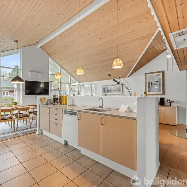 Køkken i moderne stil med trækøkkenskabe og stålarmatur, store vinduer, spiseplads, parketgulv og skråt loft. Rummelige omgivelser med naturligt lysindfald og trædetaljer.