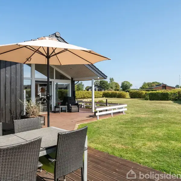 Et moderne sommerhus med lyse paneler og store vinduer står i en have. En terrasse med sorte kurvemøbler og en stor parasol tilbyder udendørs siddepladser. Græsplæne omkranser området.