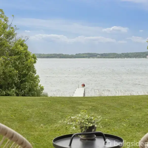 Terrasse med to kurvestole og et bord, stående på en græsplæne ved en sø. En bådebro strækker sig mod vandet, mens træer omgiver området under en blå himmel.