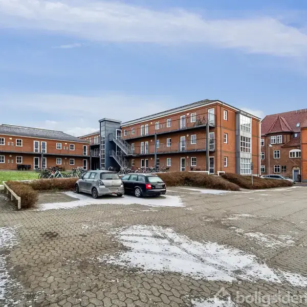 Bygning i mursten med udvendig trappe i centrum omkranset af parkeringsplads med to biler, snepletter på jorden og flere bygninger i baggrunden under en blå himmel.