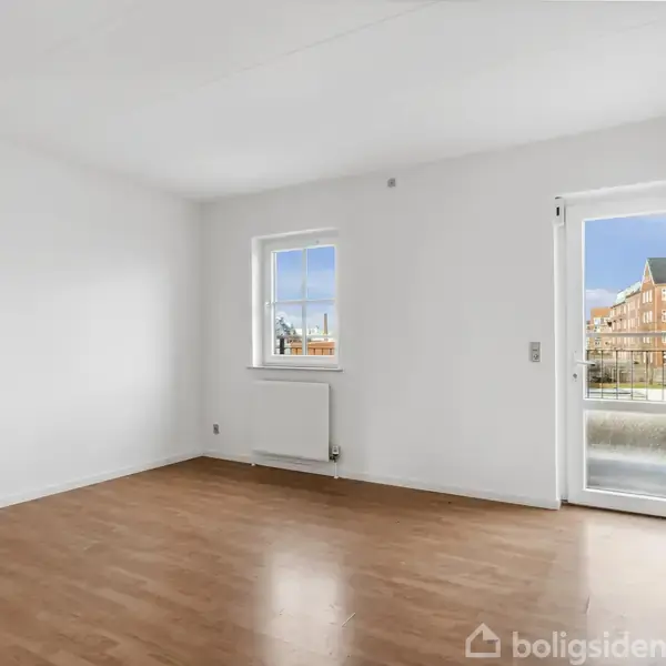 Værelse med trægulv har et stort vindue og en glasdør, der åbner ud til en balkon. Udenfor ses bygninger i et urbant område under en blå himmel.