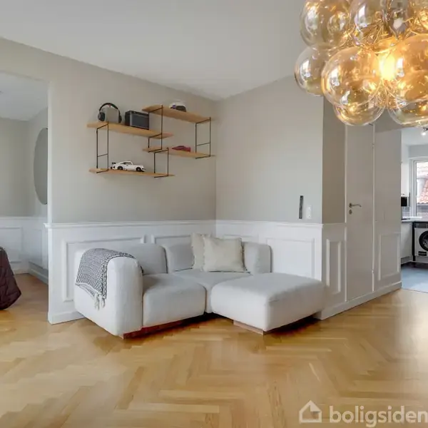 En hvid sofa står i en lys stue med parketgulv, ved siden af en moderne rund lampe. I baggrunden ses et soveværelse med teksten "GOOD MORNING SUNSHINE" på væggen.