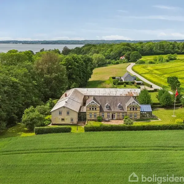 En stor gårdbygning står stille omgivet af frodige grønne marker og tæt på en skov. I baggrunden ses en sø, og en vej snor sig gennem landskabet.