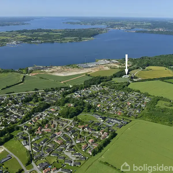Byområde ligger langs en bugt med spredte huse og grønne marker i forgrunden. En stor vandmasse strækker sig mod horisonten under en klar blå himmel.