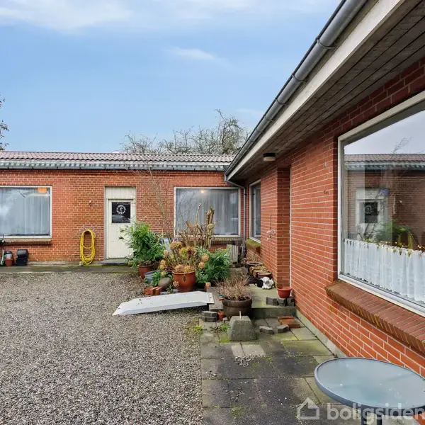 En rød murstensbygning omgiver en grusbelagt gårdsplads. Vinduerne har gardiner på indersiden, og der er spredte planter og potter i gården. En slange hænger ved døren.