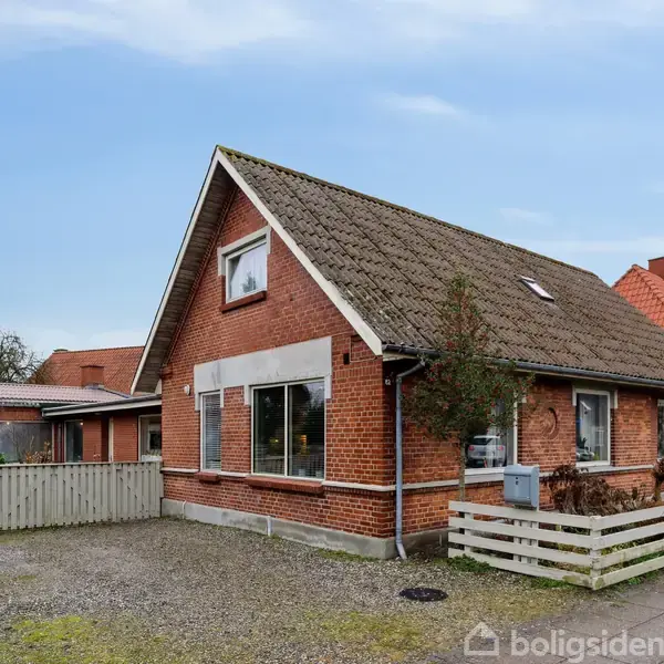 Et rødt murstenshus med sadeltag står stille langs en grusvej. Det er omgivet af et lille hegn og flere huse kan ses i baggrunden under en klar himmel.