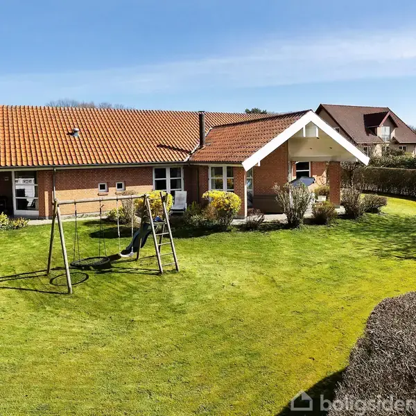 Hus med rødt tegltag står stille i en velplejet, grøn have med gyngestativ. Omgivet af hække og nabohuse under en blå himmel.