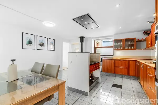 Spisebord med stole står i et lyst køkken med træskabe. Et fritstående ildsted adskiller spise- og køkkenområderne. Tre billeder hænger på væggen. Gulvet er flisebelagt.