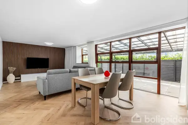 Stue med grå sofa og træspisebord står foran store vinduer, der åbner ud til en overdækket terrasse. En vægmonteret TV-skærm hænger på træpanelvæggen.