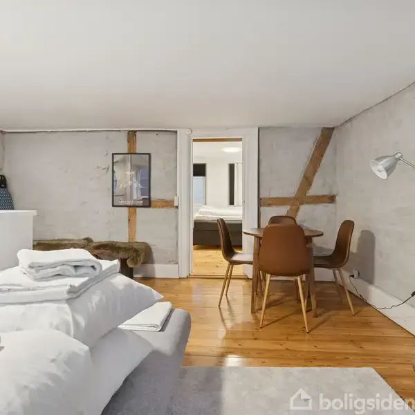 Et indendørs værelse med en hvid seng dækket af puder og håndklæder, stående på et trægulv. Ved siden af er der et rundt spisebord med stole. En gulvlampe står til højre.