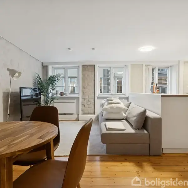 Rundt spisebord med stole står i en stue med grå sofa. Sofaen er placeret foran en væg med vinduer. I rummet ses også et TV og et åbent køkken.