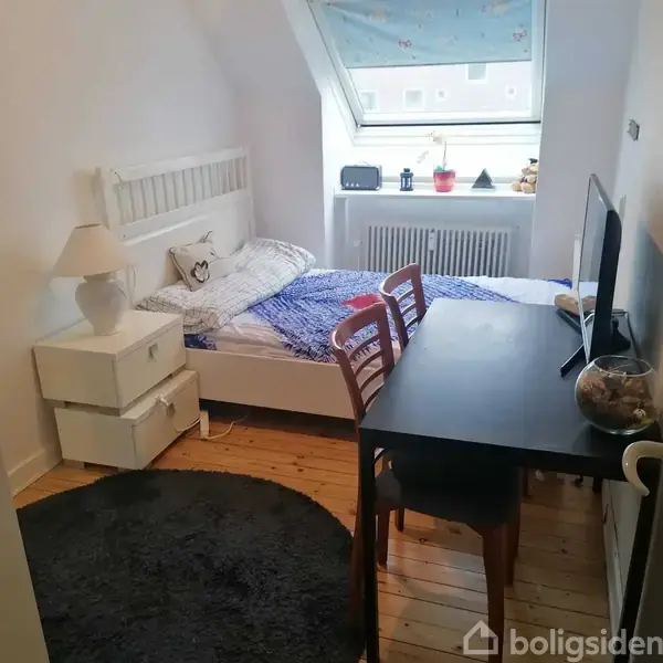 Et soveværelse har en seng med hvidt sengetøj og blå plaid, placeret ved en hvid væg. Ved siden af står et natbord med lampe. Et sort bord med stole står tæt på sengen.