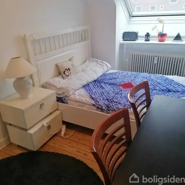 En hvid seng med en blomstret pude og blåt tæppe står i et rum med trægulv. Ved siden af sengen er der et hvidt natbord med en lampe.