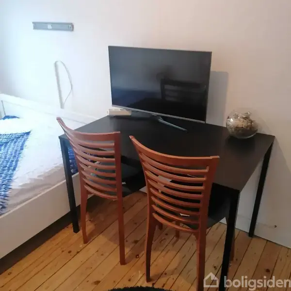 Et sort skrivebord har en fladskærms-TV på toppen, flankeret af to træstole. Ved siden af står en seng med et blåt og hvidt tæppe.