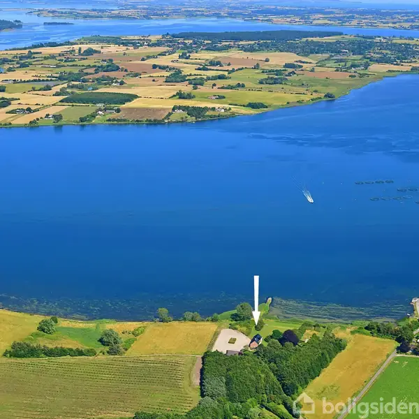 En gård omgivet af skov ligger ved en stor, blå sø. En båd sejler på søen, og i baggrunden ses spredte marker og øer under en klar himmel.