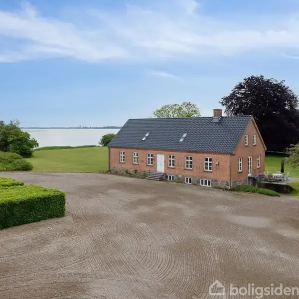 Murstensbygning med sort tag ligger stille i et åbent landskab nær en sø. En flagstang står til venstre, omgivet af grøn bevoksning. Himlen er klar med få skyer.