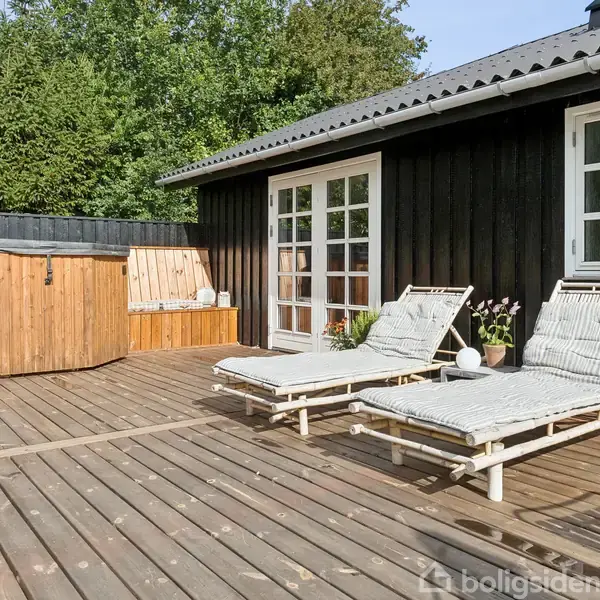 En træterrasse med to hvide liggestole opstillet foran et sortmalet træhus. En rund, træbeklædt jacuzzi står til venstre. Omgivet af grønne træer.