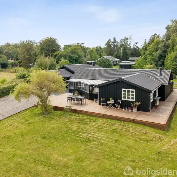 Et sort træhus med en stor træterrasse står i en frodig have med græsplæne. Omkring huset er der træer og buske, mens der ses en grusvej til venstre.
