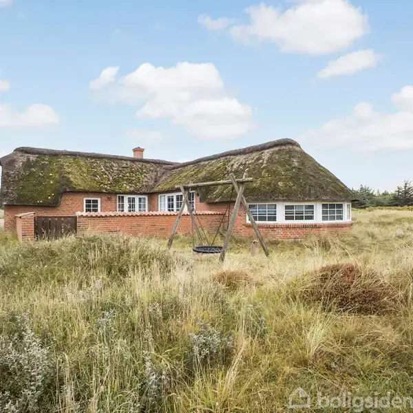 Et traditionelt, stråtækt hus står stille i en naturlig, græsbevokset eng. I forgrunden ses en enkel gynge. Himlen er blå med spredte skyer.