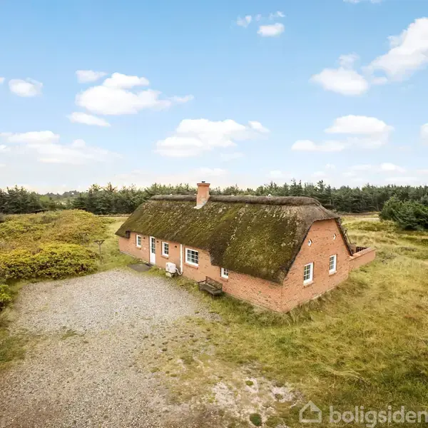 Et rødt murstenshus med stråtag står stille på en åben græsklædt grund omgivet af buske og træer under en blå himmel med spredte skyer.