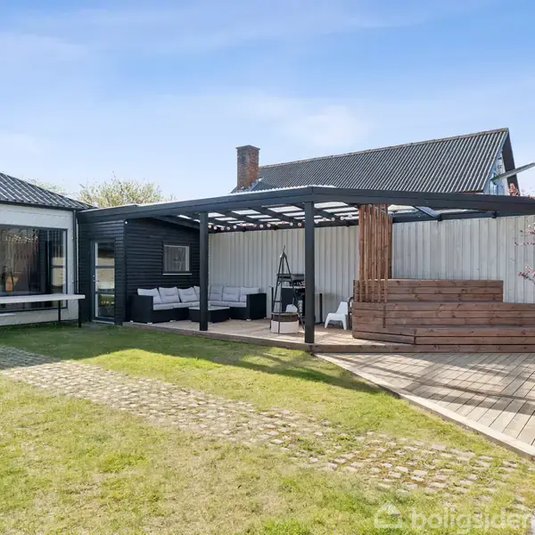 Terrasse og have i moderne stil med trædæk og pergola. Fliser og græs omkranser området. Sofaarrangement og et blomstrende træ ses i baggrunden ved siden af en hvid og sort bygning.
