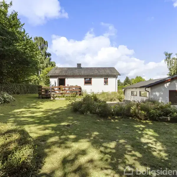Hus set fra haven, med græsplæne og buske foran. Et træskur står til højre. Himlen er blå med enkelte skyer.