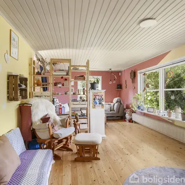 En lys stue med træloft, vægge malet i gule og rosa nuancer, og trægulv. En beige sofa og stol står foran en reol. Stort vindue med planter giver udsigt til have.
