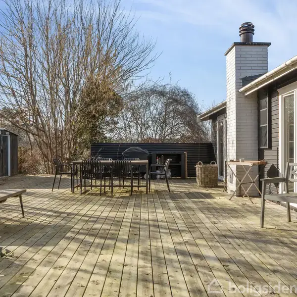 Terrasse med træmøbler står på et stort, åbent terrasseområde. Til højre er et sort træhus med store vinduespartier. Baggrunden har træer og et skur, der skaber en hyggelig atmosfære.