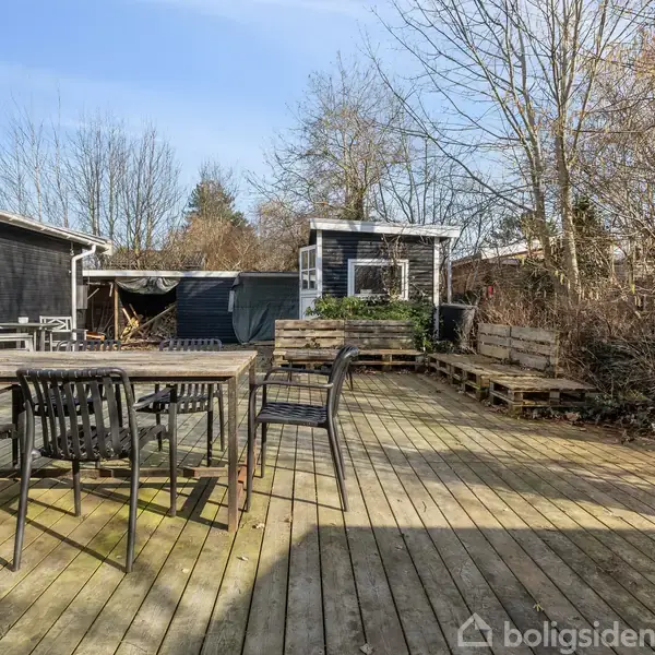 Terrasse med sort havebord og stole står stille. Omkring er der trægulv, et sort skur og omgivet af træer og buske, skaber en rolig udendørs atmosfære.