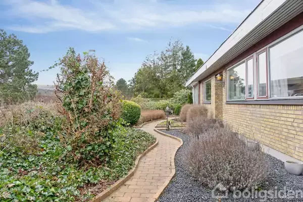 En brostenssti snor sig langs et hus med gule mursten. Omkring stien vokser buske og lavendel. I baggrunden ses en have med træer og blå himmel.