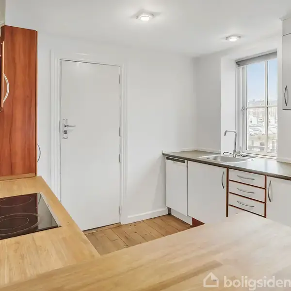 Et moderne køkken med træbordplader og glaskeramisk kogeplade, hvor lyse skabe og skuffer dominerer. En hvid dør og lille vindue skaber lysindfald i det minimalistiske rum.