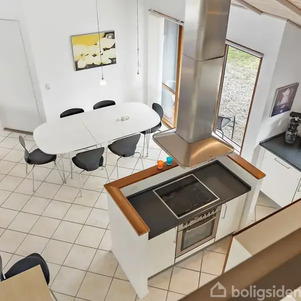 Et moderne køkken set ovenfra med en central ovn og emhætte. Til højre er der en køkkenvask og kaffemaskine. En spisebord med seks stole står tæt ved vinduet.