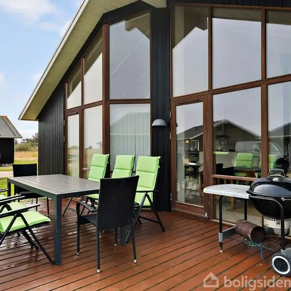 Terrasse med bord og otte grønne stole, placeres tæt på en moderne sort husfacade med store vinduer. En grill står ved siden af bordet. Det er lyst og åbent.