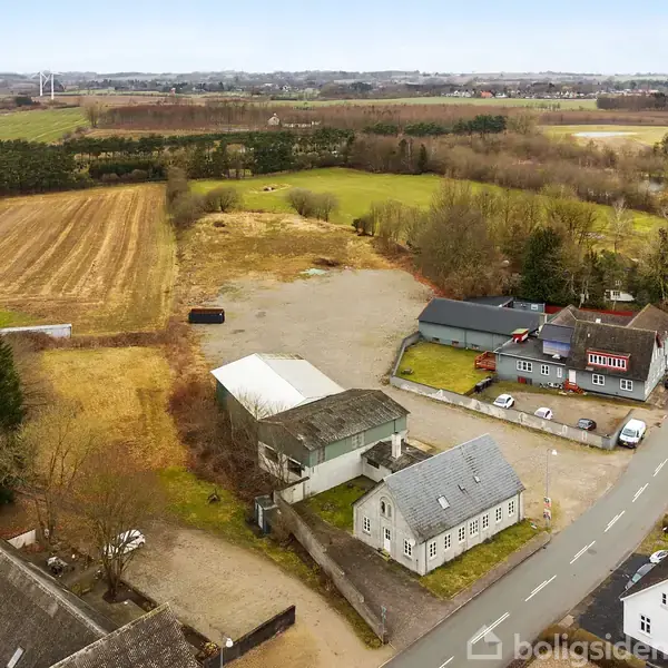 Landsbyluftfoto med landbrugsbygninger og marker. Huse grænser op til en vej, omgivet af åbne marker og skov. Vindmøller ses i baggrunden, under en overskyet himmel.