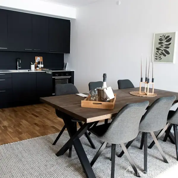 Et spisebord med seks grå stole står på et tæppe. Bordet har en bakke med forskellige genstande og lysestager. I baggrunden er der et køkken med sorte skabe.
