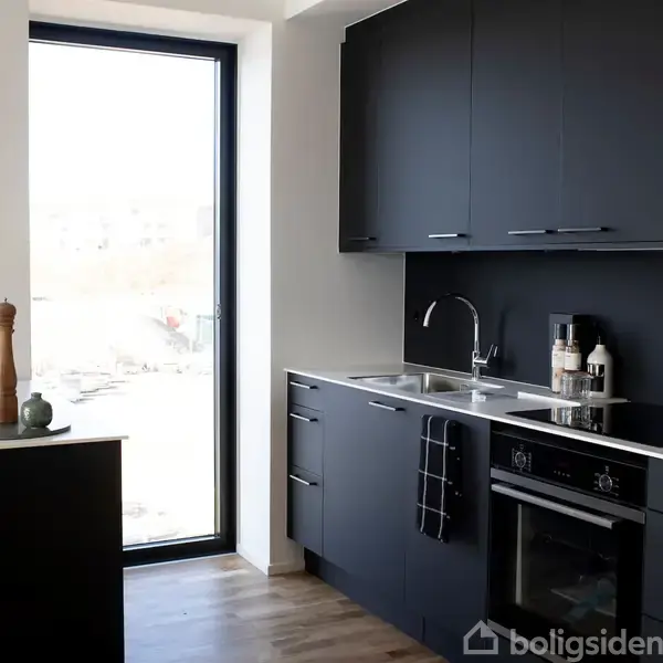 Køkken med sorte skabe og indbygget ovn, indeholdende en vask og køkkenredskaber på bordpladen. Ved siden af er et vindue, der giver lys til rummet.