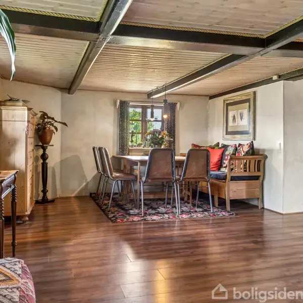 Et spisebord med seks stole står på et tæppe. I baggrunden ses et vindue og en sofa med puder. Rummet har træloft og trægulv, omgivet af planter og dekorationer.