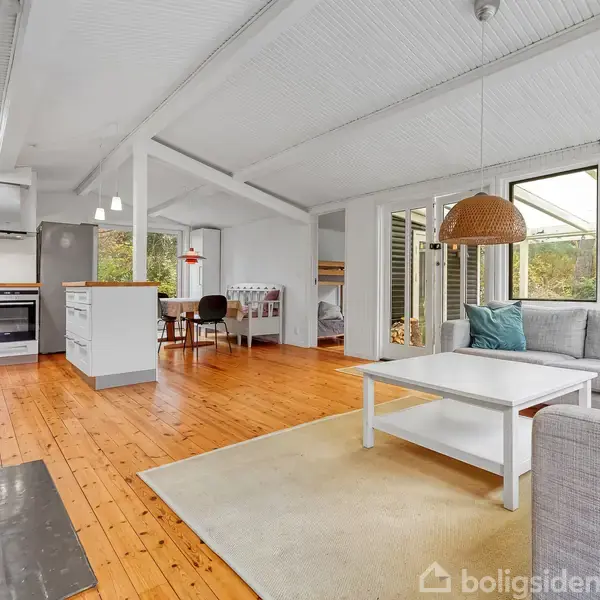Stue-alrum med gråt hjørnesofa, hvidt sofabord og gulvtæppe på trægulv, åbner op til køkken med spisekrog og store vinduer, der slipper naturligt lys ind.