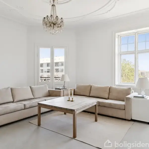 To beige sofaer står i en lys stue med et træbord imellem dem. Loftet har en dekorativ lysekrone, og store vinduer giver udsigt til bygninger udenfor.