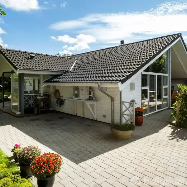 Et moderne, hvidt hus med store vinduespartier og sort tag står i en have med blomsterkrukker og en brolagt terrasse. Klare blå himmel og træer omgiver huset.